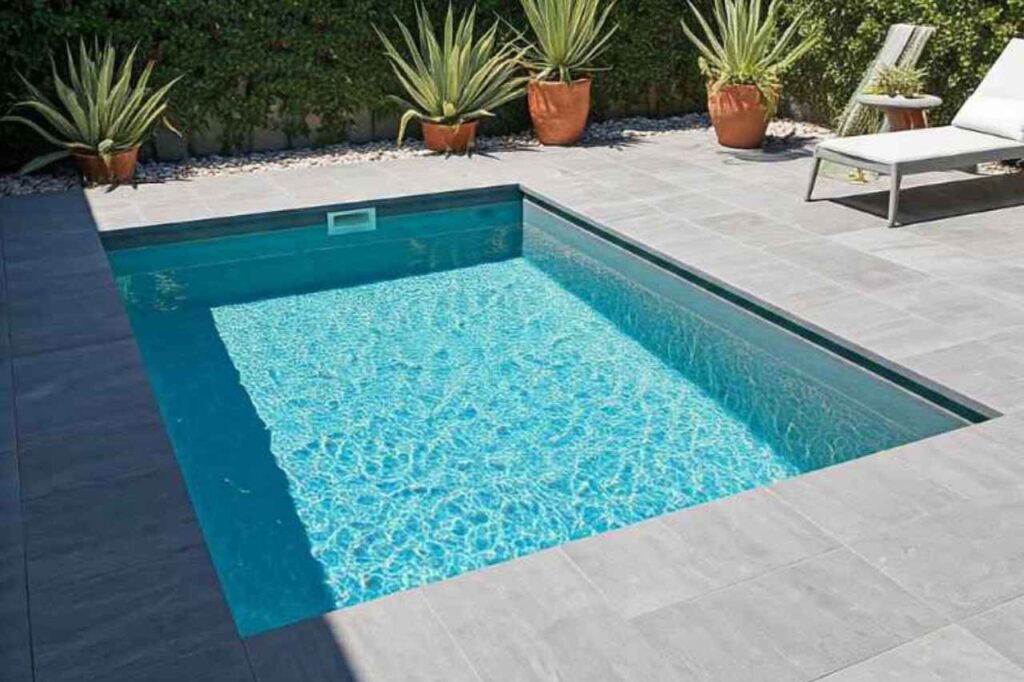A modern rectangular concrete inground plunge pool with clear blue water, surrounded by grey tiles and outdoor plants in terracotta pots.
