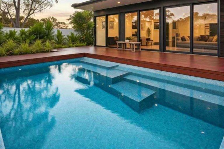 Modern backyard with a rectangular concrete plunge pool featuring wide built-in entry steps, surrounded by timber decking, lush tropical plants, and a contemporary home with large sliding glass doors glowing warmly at sunset.